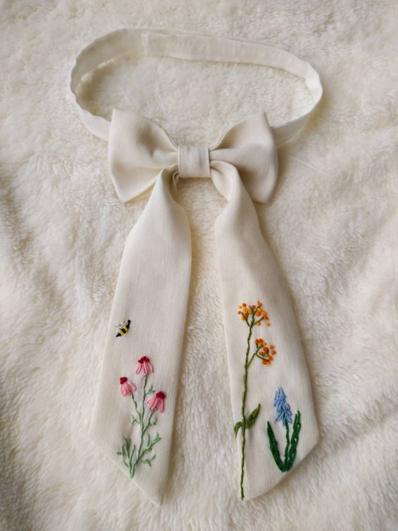 May include: A white linen bow tie with embroidered flowers and a bee. The flowers are pink, orange, blue, and green.