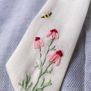 May include: White fabric with embroidered pink flowers and green leaves. A small yellow and black bee is embroidered in the top right corner.