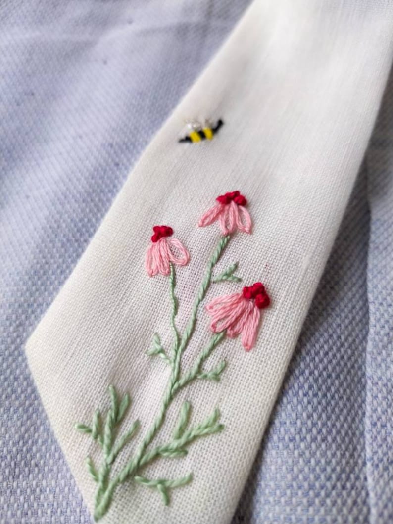 May include: White fabric with embroidered pink flowers and green leaves. A small yellow and black bee is embroidered in the top right corner.