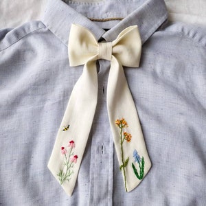 May include: A white bow tie with embroidered flowers and a bee on a light blue shirt.