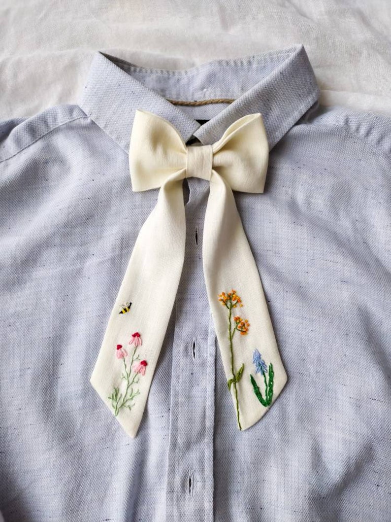 May include: A white bow tie with embroidered flowers and a bee on a light blue shirt.