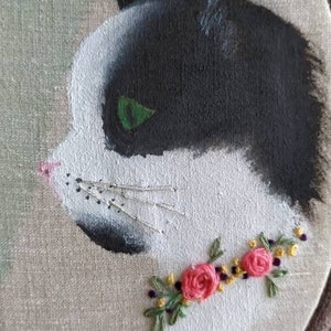 May include: Embroidered hoop art featuring a black and white cat with green eyes. The cat is surrounded by a floral garland of pink flowers and green leaves.