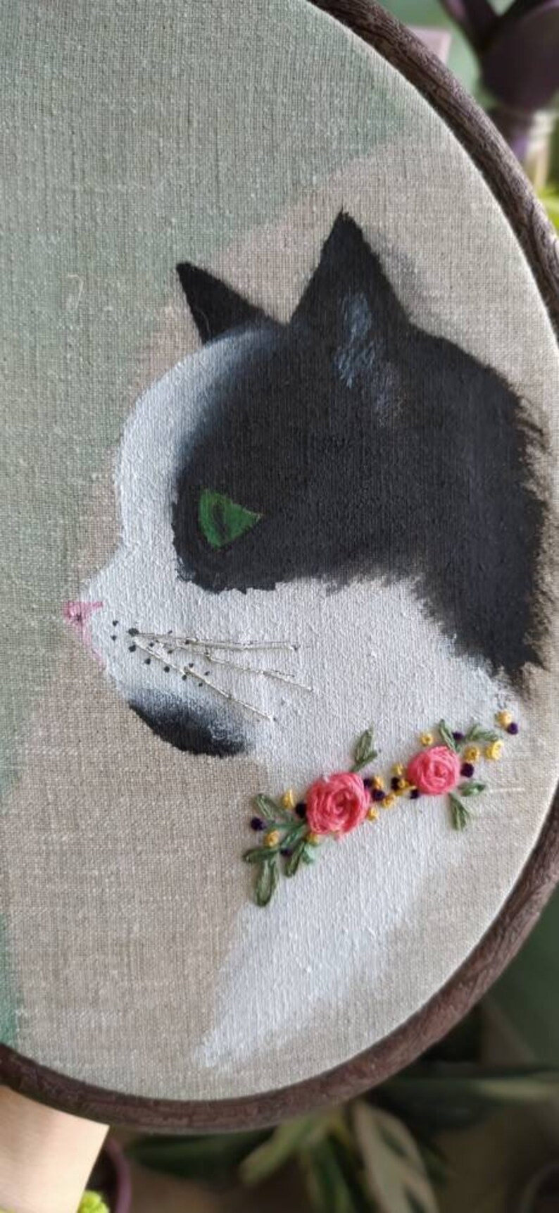 May include: Embroidered hoop art featuring a black and white cat with green eyes. The cat is surrounded by a floral garland of pink flowers and green leaves.