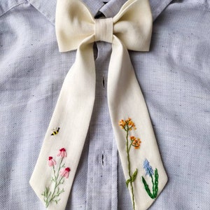 May include: A white bow tie with embroidered flowers and a bee. The bow tie is tied around a light blue shirt.