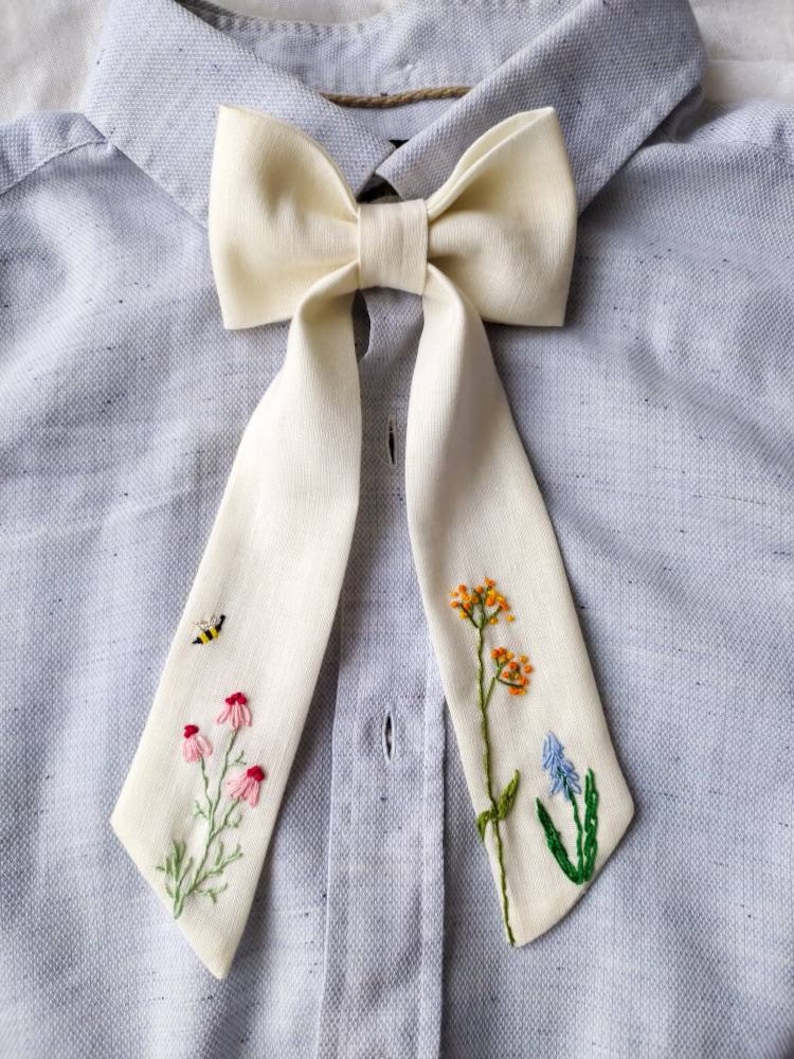May include: A white bow tie with embroidered flowers and a bee. The bow tie is tied around a light blue shirt.
