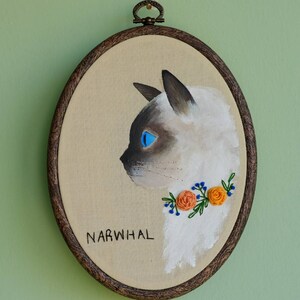 May include: Embroidered portrait of a white cat with blue eyes, wearing a floral collar with orange flowers and green leaves. The embroidery is framed in a brown wooden hoop and the word "NARWHAL" is written below the cat.