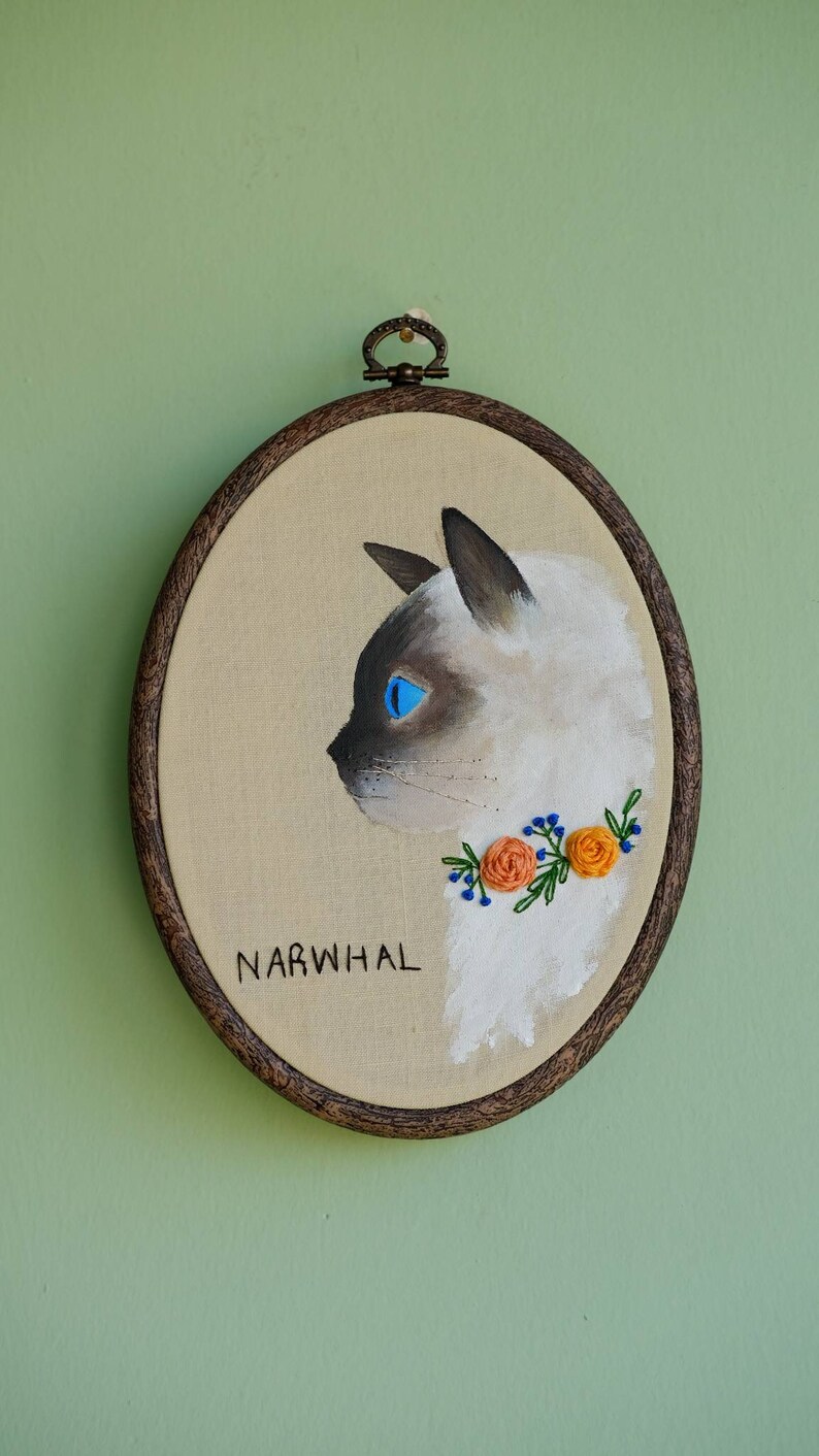 May include: Embroidered portrait of a white cat with blue eyes, wearing a floral collar with orange flowers and green leaves. The embroidery is framed in a brown wooden hoop and the word "NARWHAL" is written below the cat.