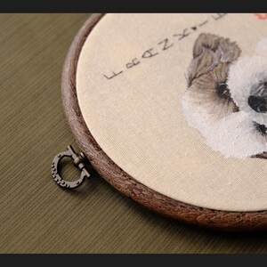 May include: Embroidered portrait of a white dog with brown markings in a wooden embroidery hoop. The hoop has a metal clasp and the fabric is beige. The text "Forever" is embroidered above the dog.