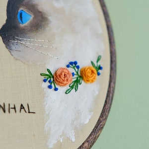 May include: Embroidered hoop art featuring a white cat with blue eyes and a floral garland of orange and blue flowers. The hoop is made of wood and has a brown stain. The text "NHAL" is embroidered below the cat.