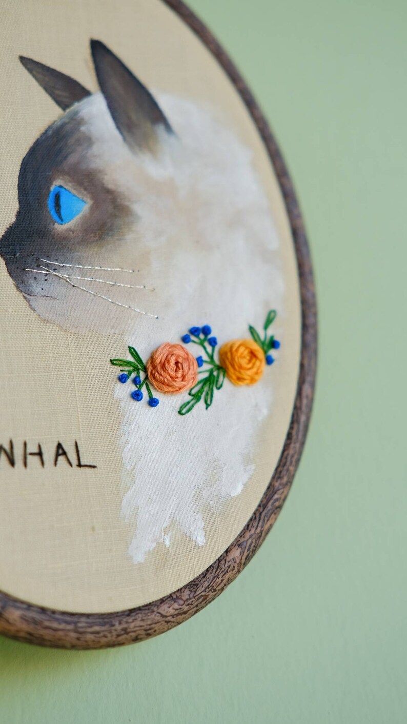 May include: Embroidered hoop art featuring a white cat with blue eyes and a floral garland of orange and blue flowers. The hoop is made of wood and has a brown stain. The text "NHAL" is embroidered below the cat.