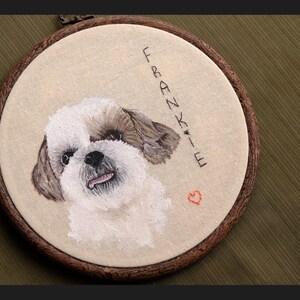 May include: Embroidered portrait of a white and brown dog with a pink nose and tongue sticking out. The embroidery is framed in a brown wooden hoop with the name "Frankie" and a small red heart embroidered below.