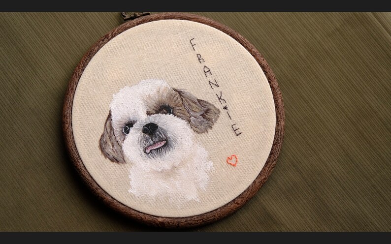 May include: Embroidered portrait of a white and brown dog with a pink nose and tongue sticking out. The embroidery is framed in a brown wooden hoop with the name "Frankie" and a small red heart embroidered below.