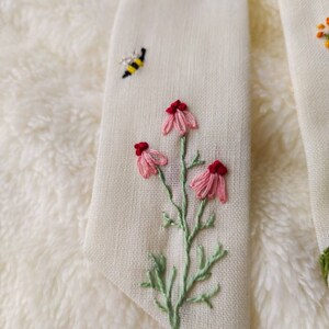 May include: A white fabric with embroidered pink flowers, green leaves, and a yellow and black bee.