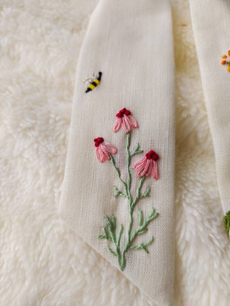 May include: A white fabric with embroidered pink flowers, green leaves, and a yellow and black bee.