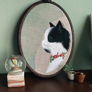 May include: A black and white cat with green eyes embroidered on a beige fabric in a round wooden embroidery hoop. The cat is wearing a red flower garland.