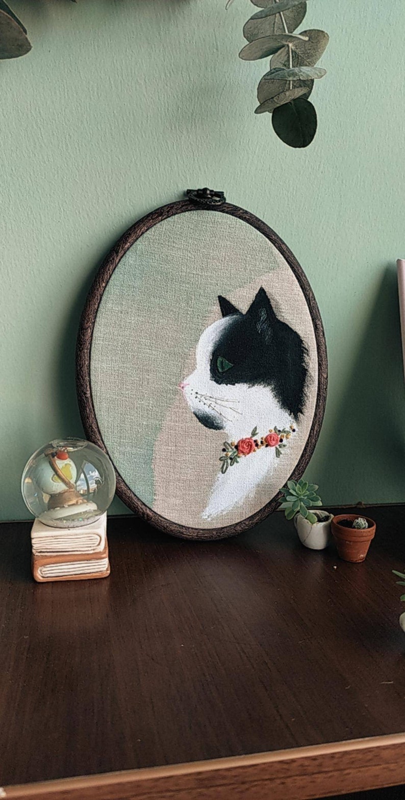 May include: A black and white cat with green eyes embroidered on a beige fabric in a round wooden embroidery hoop. The cat is wearing a red flower garland.