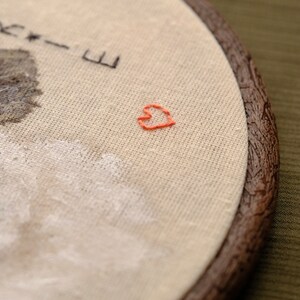 May include: Embroidered hoop art with a brown wooden frame. The fabric is beige and features a small, hand-stitched red heart in the center. The text "Nikki-E" is embroidered in black thread at the top of the hoop.