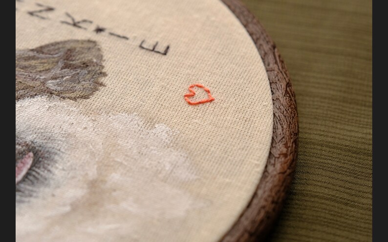 May include: Embroidered hoop art with a brown wooden frame. The fabric is beige and features a small, hand-stitched red heart in the center. The text "Nikki-E" is embroidered in black thread at the top of the hoop.