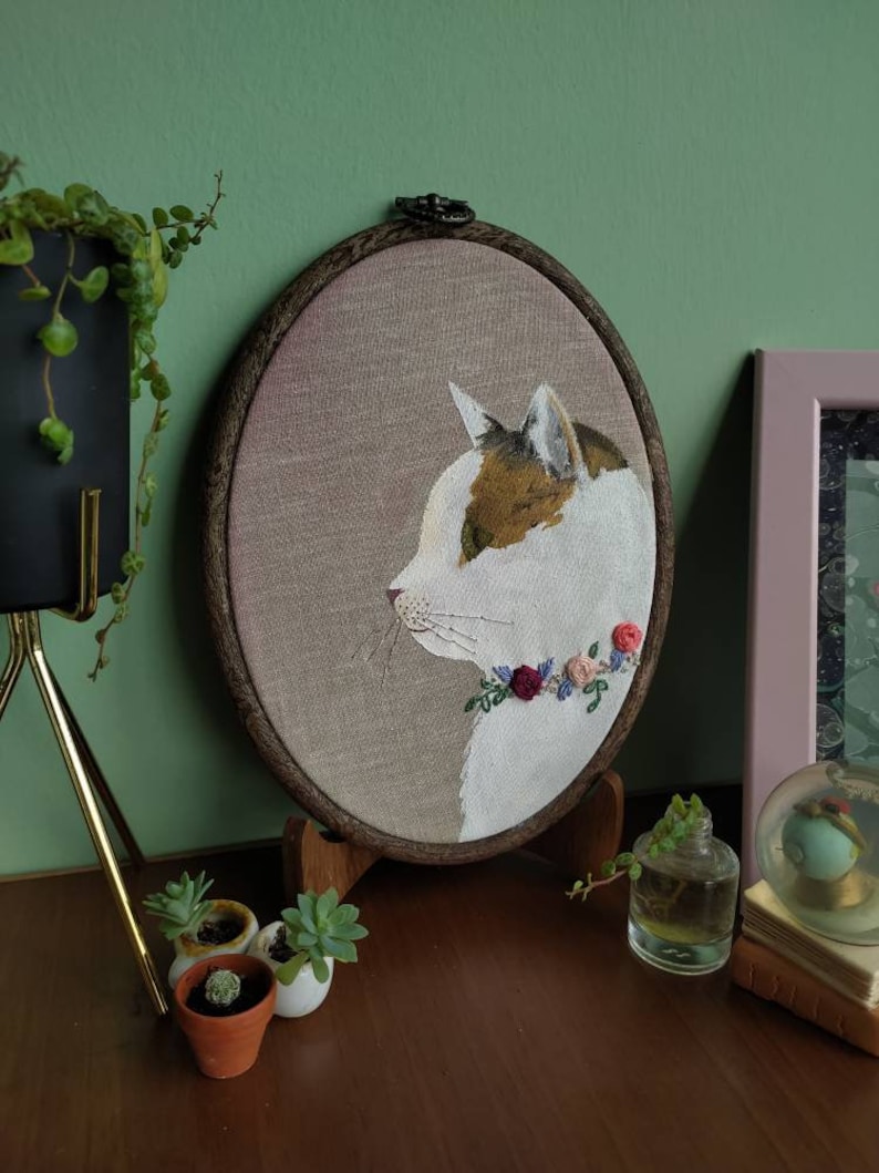 May include: Embroidered portrait of a white cat with brown and orange markings, wearing a floral wreath around its neck. The embroidery is framed in a wooden hoop and displayed on a wooden stand.