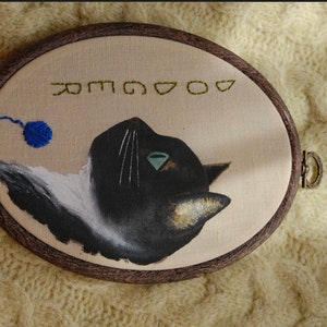 May include: Embroidered hoop art featuring a black and white cat with a green eye, looking to the right. A blue ball of yarn is in the upper left corner. The text "GOOD" is embroidered in green above the cat.