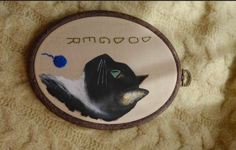 May include: Embroidered hoop art featuring a black and white cat with a green eye, looking to the right. A blue ball of yarn is in the upper left corner. The text "GOOD" is embroidered in green above the cat.