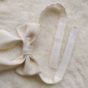 May include: A white fabric bow tie with a long, adjustable strap. The strap is made of a white fabric with a hook and loop closure.