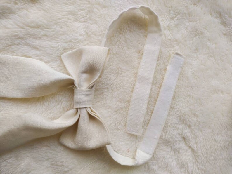 May include: A white fabric bow tie with a long, adjustable strap. The strap is made of a white fabric with a hook and loop closure.