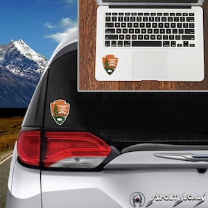 May include: A white car with a National Park Service sticker on the back window. The sticker is a brown and green shield with a mountain range, a tree, and a bison inside. The car is parked in front of a mountain range.