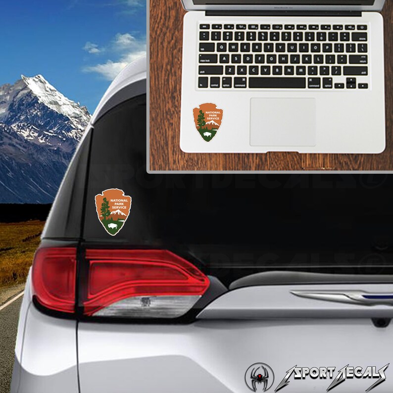May include: A white car with a National Park Service sticker on the back window. The sticker is a brown and green shield with a mountain range, a tree, and a bison inside. The car is parked in front of a mountain range.