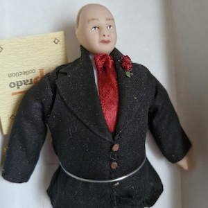 May include: A porcelain doll dressed in a black suit with a red tie and a small red flower on the lapel. The doll has a bald head and a friendly expression.