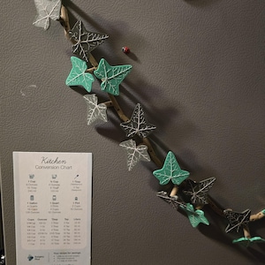 May include: A kitchen conversion chart magnet with a green ivy vine design. The chart includes measurements for cups, ounces, tablespoons, teaspoons, gallons, quarts, pints, and liters. The chart also includes a phone number for a company called "Surgery Plus".