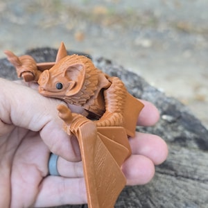 May include: A brown articulated bat figurine featuring a detailed head with pointed ears, a snout, and black eyes. The figure has textured wings with visible joints, allowing for movement, and a molded body with fine fur-like detailing.