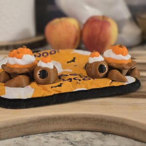 May include: Three brown, eye-shaped Halloween-themed decorations with pumpkin and whipped cream toppers sit on a yellow and black patterned mat. Two apples are in the background. The decorations are on a wooden cutting board.