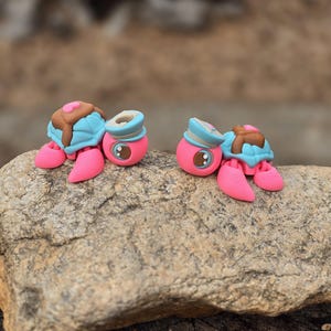 May include: Two whimsical, handmade turtle figurines. Each turtle has a pink body, pink feet, and a blue shell topped with brown frosting and a pink heart. The turtles are displayed on a gray rock.