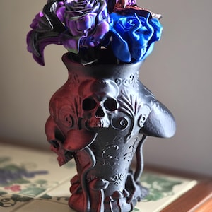 May include: A black vase with a skull design, holding a bouquet of purple and blue artificial roses. The vase has intricate details and a dark, gothic aesthetic. The roses add a pop of color to the arrangement.