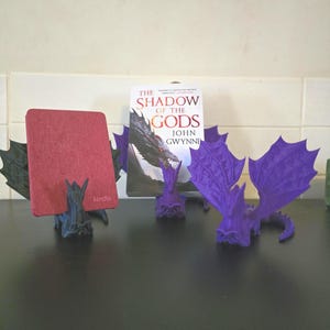 May include: Three 3D printed dragon bookends, two are purple and one is black. The black dragon is holding a red book with the word "kindle" on it. The purple dragons are holding a book titled "The Shadow of the Gods" by John Gwynne.