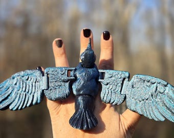 Puede incluir: Una escultura de pájaro azul oscuro con las alas extendidas, sostenida en una mano. El pájaro tiene plumas detalladas y un acabado metálico texturizado con reflejos azules. Es un objeto decorativo.