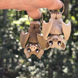 May include: Two brown and beige bat-shaped keychains with black eyes and white teeth hanging from silver key rings.