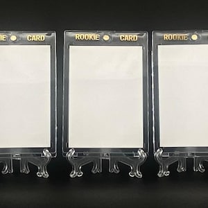 May include: Three clear acrylic card holders with stands. Each holder displays a white card and has gold lettering that reads "ROOKIE CARD". The holders are designed to display collectible cards.