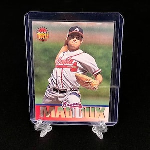 May include: A baseball trading card featuring Greg Maddux in a white and red Atlanta Braves uniform, captured in a pitching motion. The card includes the text Triple Play and Maddux at the bottom, all encased in a clear protective plastic sleeve.