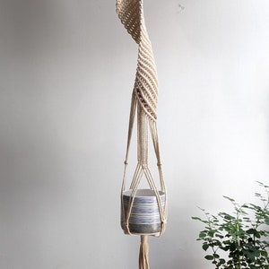 May include: A cream-coloured macramé plant hanger with a spiral design, holding a ceramic pot with blue and white stripes. The pot is suspended from a wooden ring, with a tassel at the bottom. A potted plant is in the background.