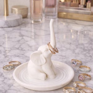 May include: A white ceramic elephant ring holder with a raised trunk, holding two gold rings. The elephant sits on a round, ridged dish, surrounded by various rings. The background includes perfume bottles and a marble surface.