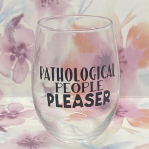 May include: A clear glass wine glass with the text "Pathological People Pleaser" printed in black on the side.