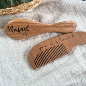 May include: Two wooden hair care items: a brush with "Rafael Berlin" engraved and a comb with "you are loved" engraved. Both are made of light-colored wood and rest on a neutral-toned fabric. The brush has a rounded handle.