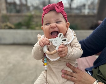 May include: A happy baby wearing a burgundy bow headband and a beige onesie. The baby is holding a wooden and silicone teether with a bunny design. A pacifier and colourful beads are attached to the teether.