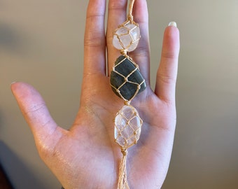 Puede incluir: Un amuleto de cristal con tres piedras, un cuarzo transparente, una piedra verde oscuro y un cuarzo blanco, envuelto en cuerda de macramé.