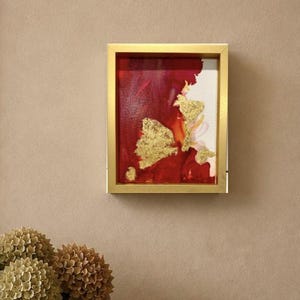 Puede incluir: Una pieza de arte mural rectangular que presenta un diseño abstracto en rojo intenso y blanco con detalles de pan de oro, encerrada en un marco de oro metálico. La obra se exhibe en una pared de tonos neutros, mostrando un contraste audaz.