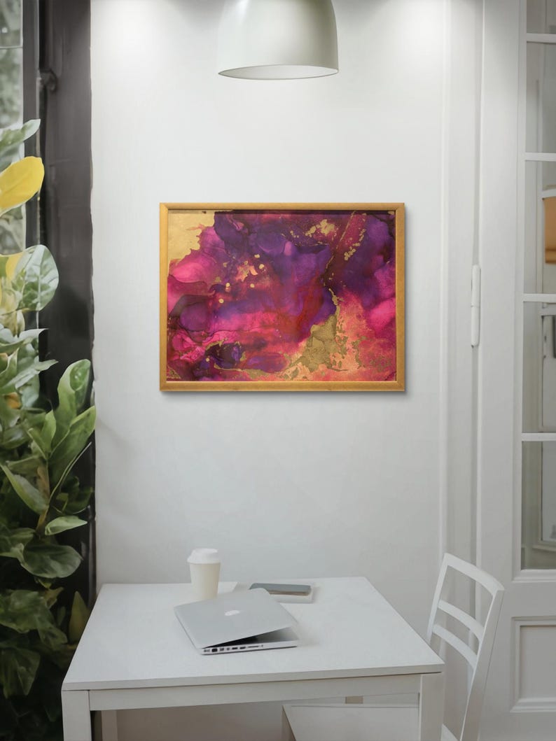 May include: Abstract painting in a wooden frame with shades of pink, purple, and gold. The painting is hanging on a white wall above a white table with a laptop and a coffee cup.