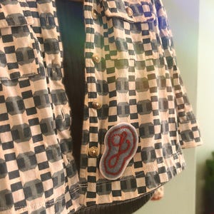 May include: A button-down shirt featuring a geometric pattern of dark gray and cream squares. The shirt includes two chest pockets, light-colored buttons, and a light blue oval patch on the lower front with a red embroidered cursive letter J.
