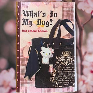 May include: A rectangular book cover titled What's In My Bag? law school edition. The cover features a black tote bag with a white Hello Kitty charm and a small doll charm, set against a pink and white plaid background with floral accents.